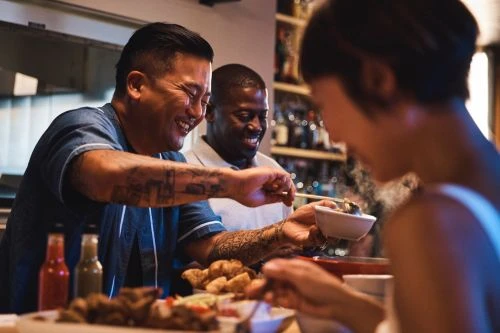 Three adults, two men and one woman, sit at a bar enjoying food and drinks in a warmly lit restaurant setting, sharing a laugh together.