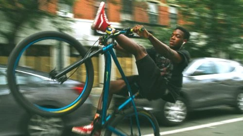 Young man performs a bike trick on a blue and yellow BMX on a city street, wearing black shorts and a t-shirt, plus red high-tops sneakers.