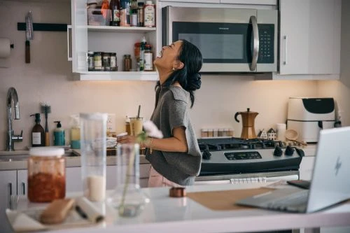 Woman laughs in a brightly lit kitchen, opening a cabinet stocked with spices and condiments; a laptop sits on the kitchen island.