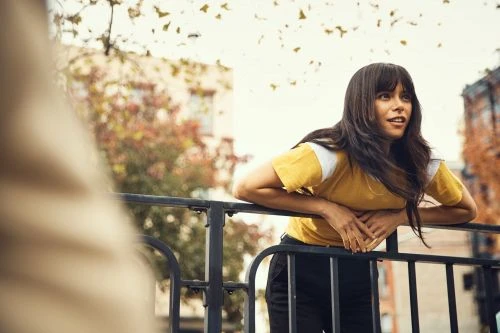 Woman with dark hair and yellow T-shirt leans on a metal railing with trees and buildings in the blurred background in daylight.