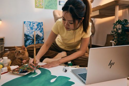 Asian woman with her cat painting a green plant on large paper with a paintbrush, while using a laptop for reference indoors.