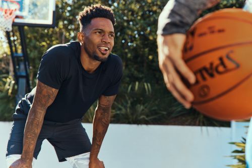 Athletic man in black t-shirt with arm tattoos pauses during an outdoor basketball game on a sunlit court in an urban setting.