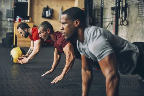 Three athletic men are doing push-ups in a gym, with a yellow medicine ball beside them, in a high-energy fitness setting.