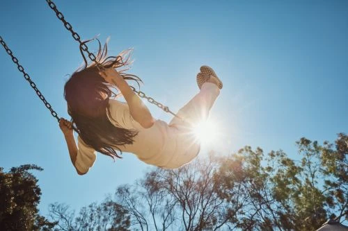 Child enjoys a sunny day swinging high in the park, with a bright sunburst adding a playful touch to the carefree scene.