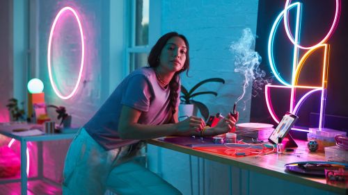 Woman with a pipe leans on a counter with a computer, art supplies, and neon lights in the background, creating a vibrant, modern atmosphere.