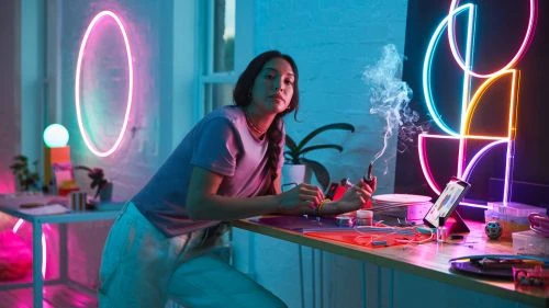 Woman with a pipe leans on a counter with a computer, art supplies, and neon lights in the background, creating a vibrant, modern atmosphere.