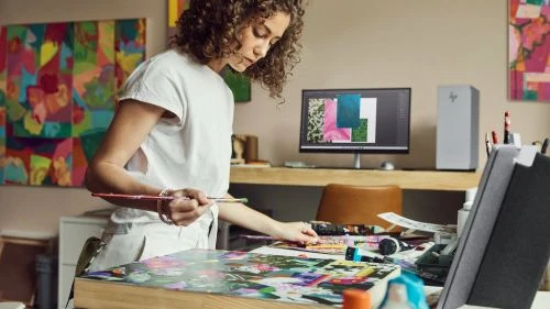 A woman artist stands at a table in a studio, painting with acrylics. A computer and desk are in the background.