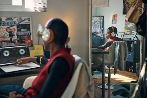 Person with headphones using a laptop at a desk in a home interior. Another person with headphones visible in a mirror reflection.