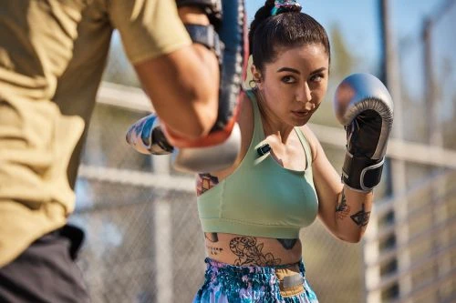 A female boxer wearing boxing gloves spars with a trainer outdoors on a sunny day, showcasing strength and determination in a dynamic setting.