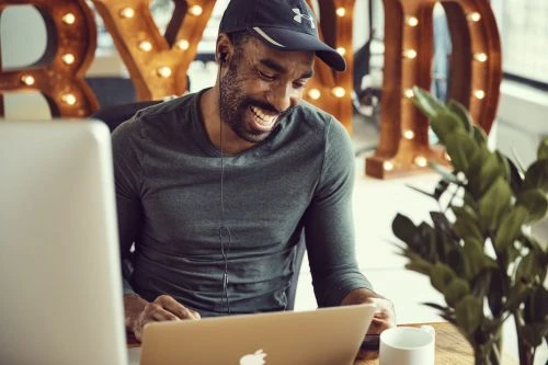 Man with a beard and baseball cap works on a laptop in an office with light-bulb-lined marquee letters and a plant.