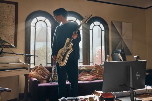 Person in black sweater holding electric guitar in a naturally lit home music studio with a piano, computer, and large arched windows.