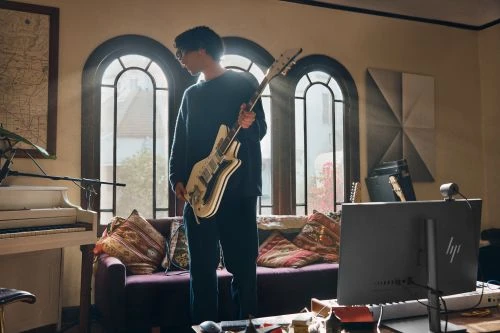 Person in black sweater holding electric guitar in a naturally lit home music studio with a piano, computer, and large arched windows.