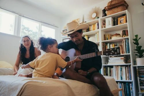 Man wearing a cowboy hat playing acoustic guitar while a child touches the strings and a woman watches from a bed