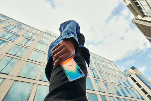 Low-angle shot of a person holding a Samsung smartphone in an urban setting with tall buildings in the background under a cloudy sky.