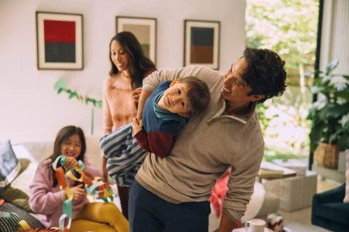 Family of four in living room: father lifting son, mother smiling, daughter playing with colorful toy. Casual indoor scene with natural light.