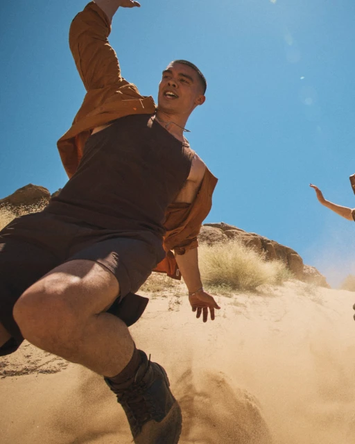 Action shot of a man jumping on a sand dune in a desert landscape on a sunny day, wearing a brown jacket and dark athletic wear.