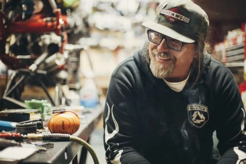 Smiling man in a cap and glasses, wearing a hoodie in a workshop setting, with tools and equipment visible in the background.