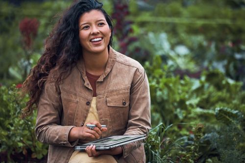 Smiling Asian woman in a field of green plants, wearing a tan jacket and holding a tablet and pen, with a shallow depth of field.