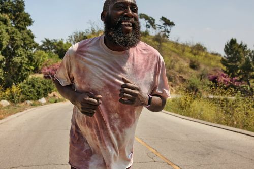 A bearded African American man runs outdoors on a sunny day, wearing a tie-dye shirt and smartwatch, with trees in the background.