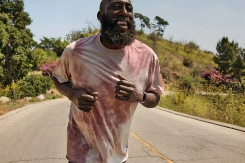 A bearded African American man runs outdoors on a sunny day, wearing a tie-dye shirt and smartwatch, with trees in the background.