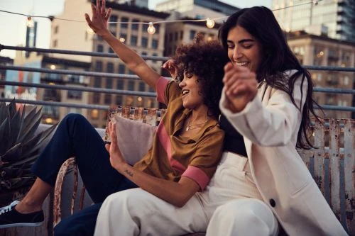 Two women laugh and dance on a rooftop at dusk, wearing casual clothing. City buildings and string lights are visible in the background.