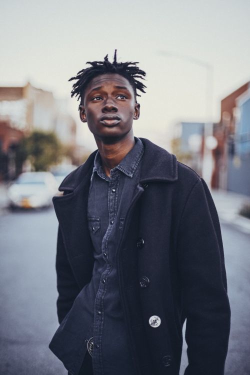 Portrait of a young black man with dreadlocks wearing a dark peacoat and denim shirt, standing on a street with buildings and a car in background.