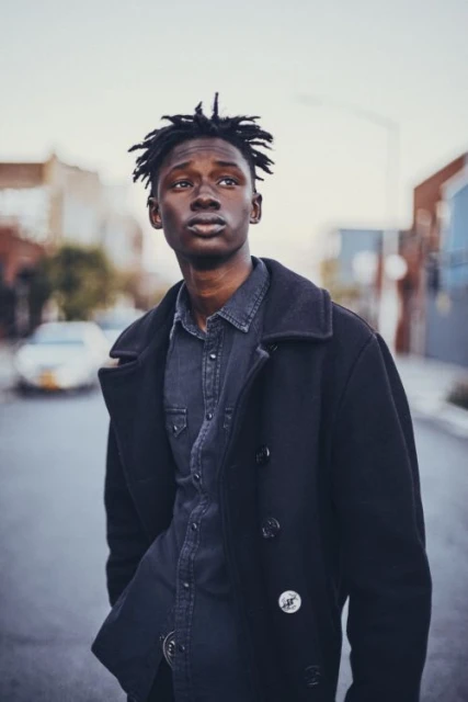 Portrait of a young black man with dreadlocks wearing a dark peacoat and denim shirt, standing on a street with buildings and a car in background.
