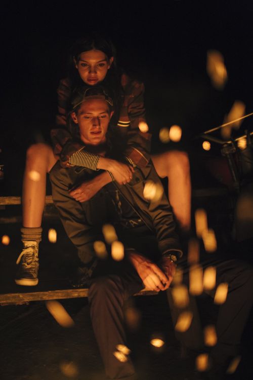 Young adult couple sits outside at night, illuminated by orange light and bokeh; woman embraces man in a nighttime outdoor setting.