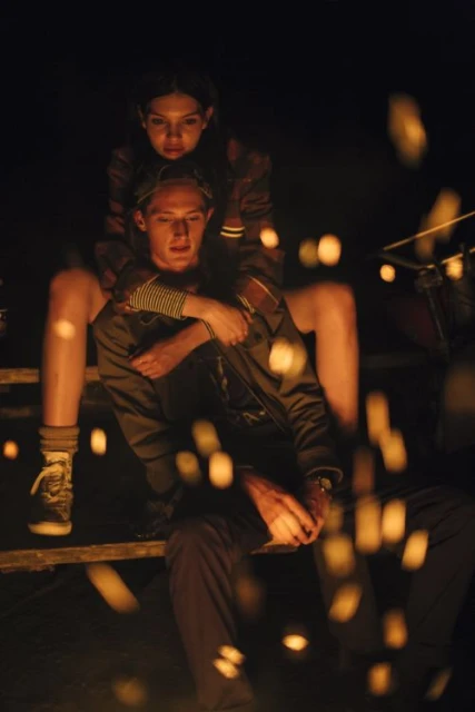 Young adult couple sits outside at night, illuminated by orange light and bokeh; woman embraces man in a nighttime outdoor setting.