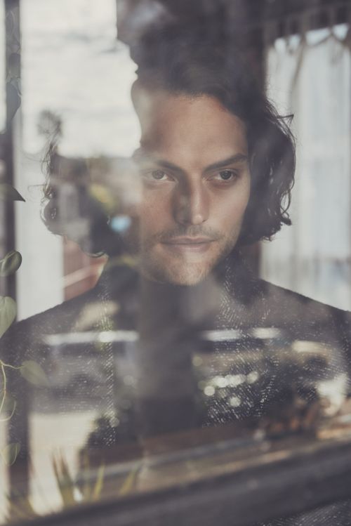 Portrait of a dark-haired adult male looking through a window with reflections of indoor plants and urban architecture in the background.