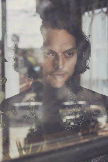 Portrait of a dark-haired adult male looking through a window with reflections of indoor plants and urban architecture in the background.
