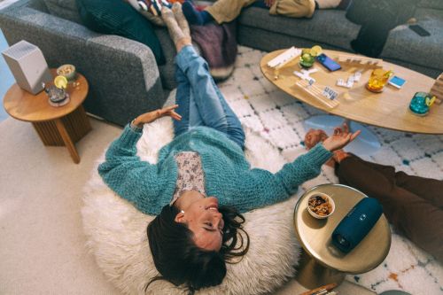 Smiling woman lies on a cushion surrounded by friends and objects, including a speaker, drinks, snacks, and a wooden game, in an indoor space.