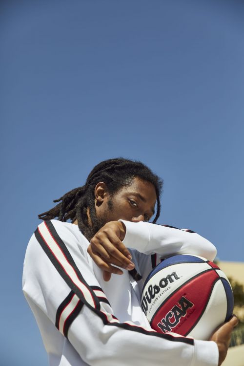 Adult male with locs holds a Wilson NCAA basketball against a sunny blue sky, wearing a white long-sleeved shirt with striped sleeves.