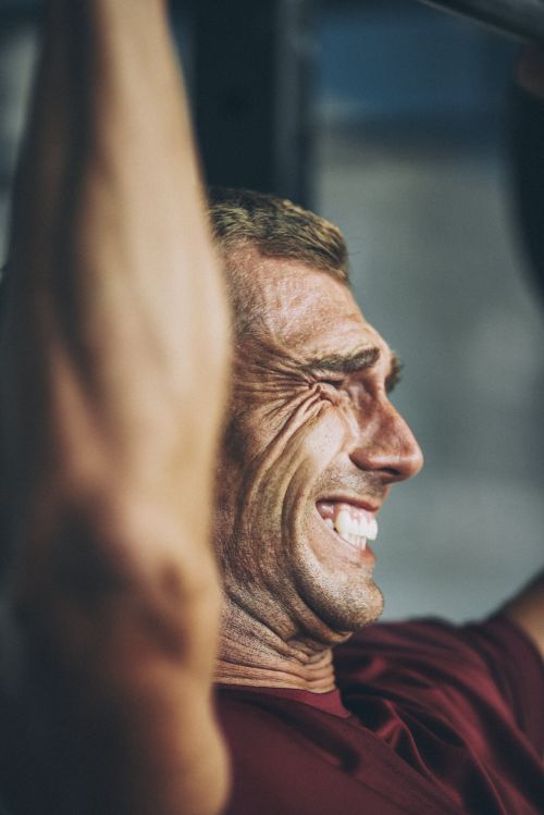 An adult male with a grimacing expression is doing pull-ups; he is wearing a red t-shirt against a gray background inside a fitness setting.