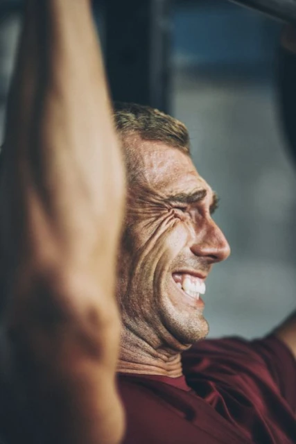 An adult male with a grimacing expression is doing pull-ups; he is wearing a red t-shirt against a gray background inside a fitness setting.