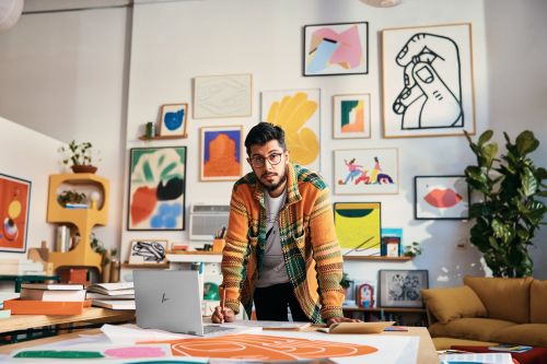 Man stands at a desk with a laptop, surrounded by colorful art in an indoor setting. He wears glasses and a patterned sweater.