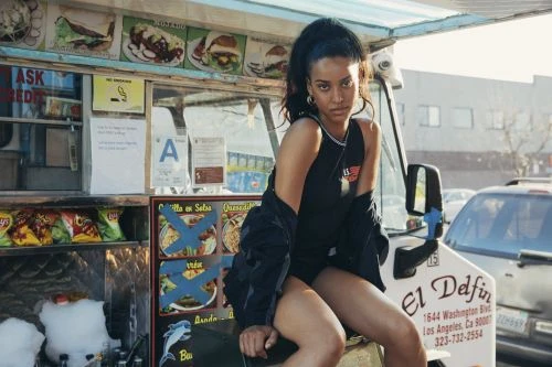 An adult woman with dark hair poses on a food truck against an urban background, showcasing modern style and casual confidence.