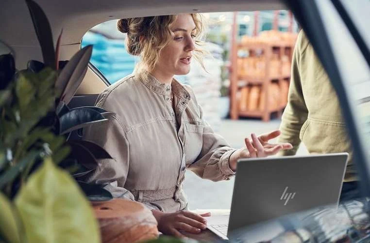 Woman working on laptop in car trunk, surrounded by potted plants; another person is seen with an open warehouse in the background.