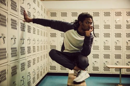 A young Black man with dreadlocks poses in a locker room wearing athletic clothing and sneakers, sitting on a bench with a locker background.