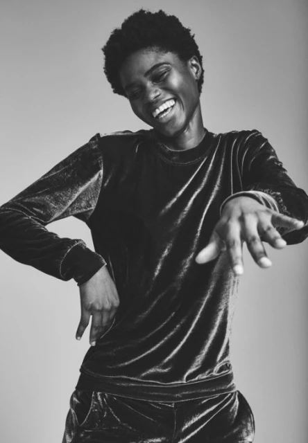 A Black woman with an expressive smile poses indoors in a black velvet top and pants, showcasing a relaxed and joyful mood.