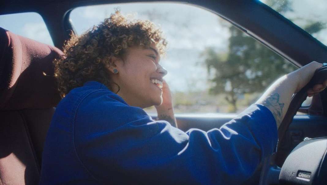 Young woman driving a car on a sunny day, smiling and enjoying the ride, with trees visible in the background, wearing a blue jacket.