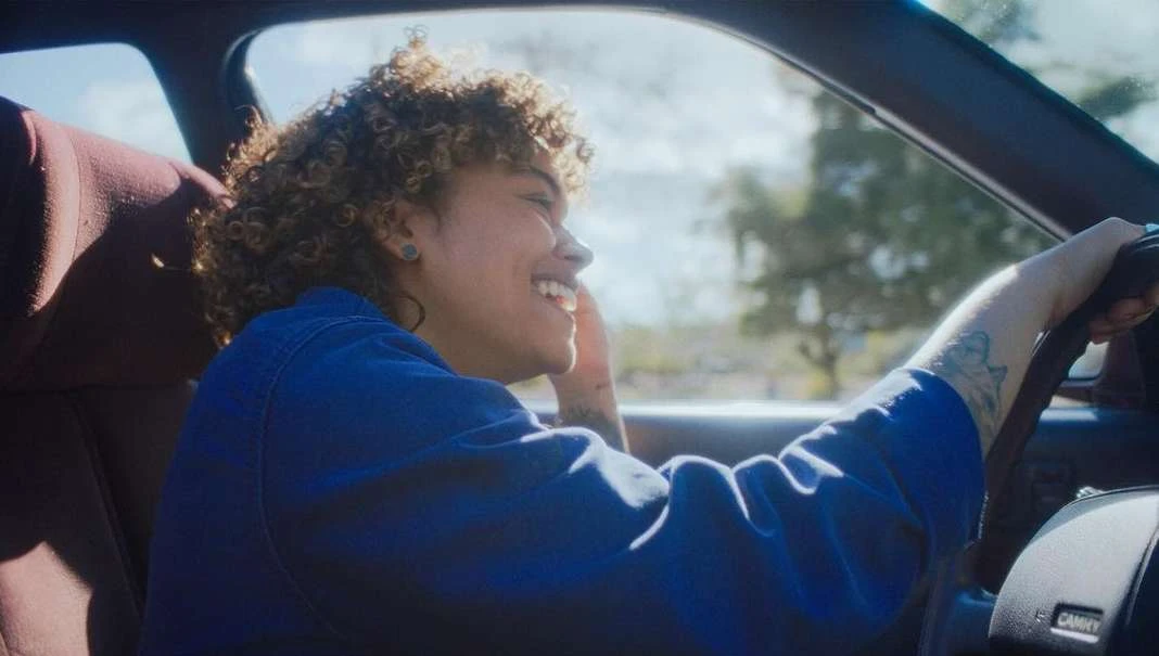 Young woman driving a car on a sunny day, smiling and enjoying the ride, with trees visible in the background, wearing a blue jacket.