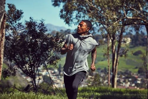 An adult male running outdoors on a sunny day wearing a sleeveless shirt and athletic leggings with trees and mountains in the background.
