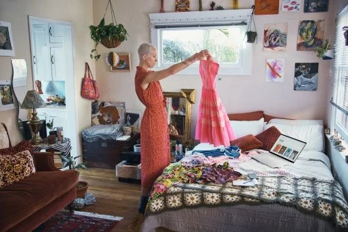 Adult female stands in sunlit room, holding up pink dress with crochet blanket and laptop visible. Wall art and plants add character.