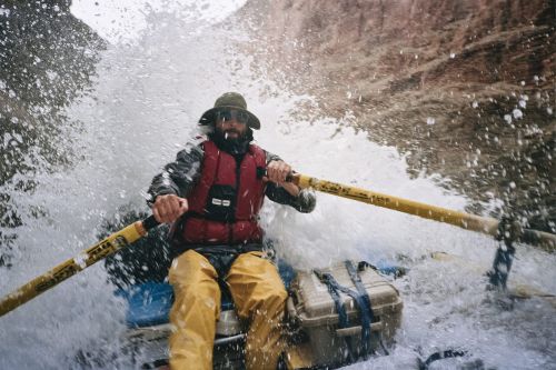 A man in a raft navigates white water rapids, wearing a life vest and hat, rowing with yellow oars amid splashing water.