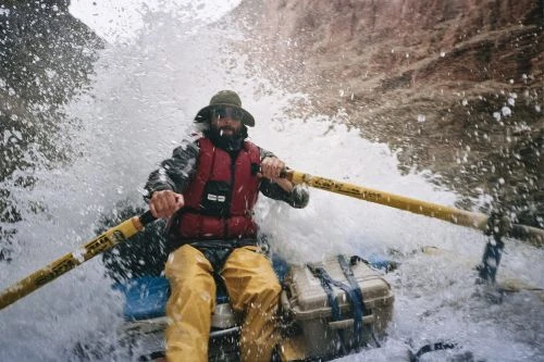 A man in a raft navigates white water rapids, wearing a life vest and hat, rowing with yellow oars amid splashing water.