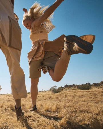 Woman jumping and laughing outdoors in a desert landscape wearing beige shorts and a knit top, photographed by Jeff Vallee