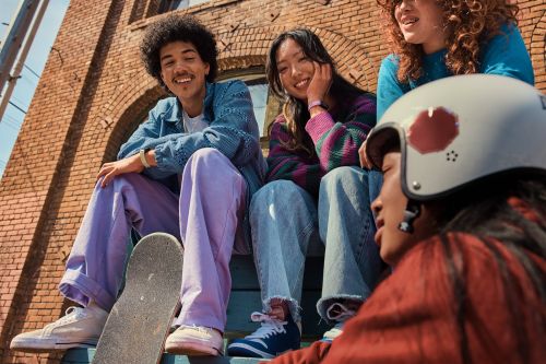 Group of four young adults sits on outdoor steps in casual clothing. Skateboard rests against steps in front of brick building.
