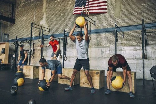 A group of athletic men in a gym perform strength and conditioning exercises using medicine balls and plyometric boxes in an indoor fitness setting.