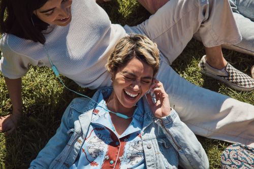 Two women relaxing on green grass, one laughing with earbuds, wearing a light blue denim jacket and printed shirt; the other with a white sweater.